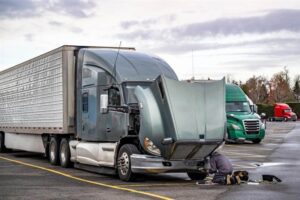 Truck driver repairing broken gray big rig semi truck with open hood.